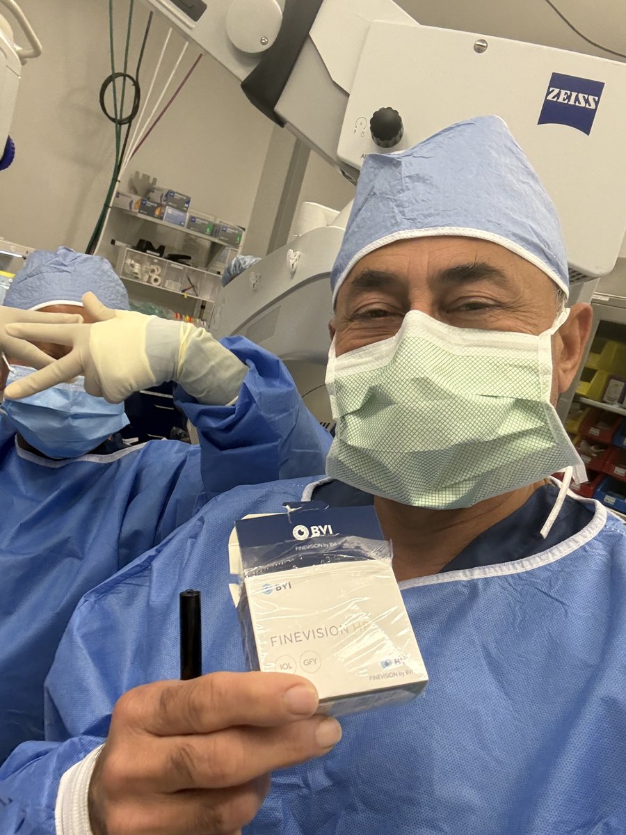 Masked surgeon in a blue cap and gown beside a ZEISS surgical microscope, holding up a boxed BVI FineVision HP lens implant