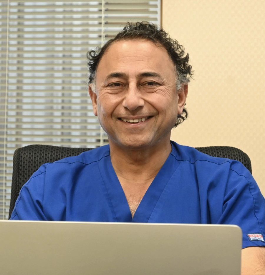 Dr. Rajesh Khanna of Khanna Vision Institute in blue scrubs, smiling at a desk with a laptop