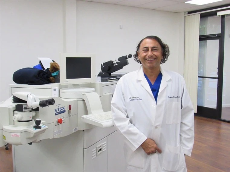 Dr. Rajesh Khanna in a white coat and blue scrubs smiling beside a VISX excimer laser in a Khanna Vision Institute procedure room