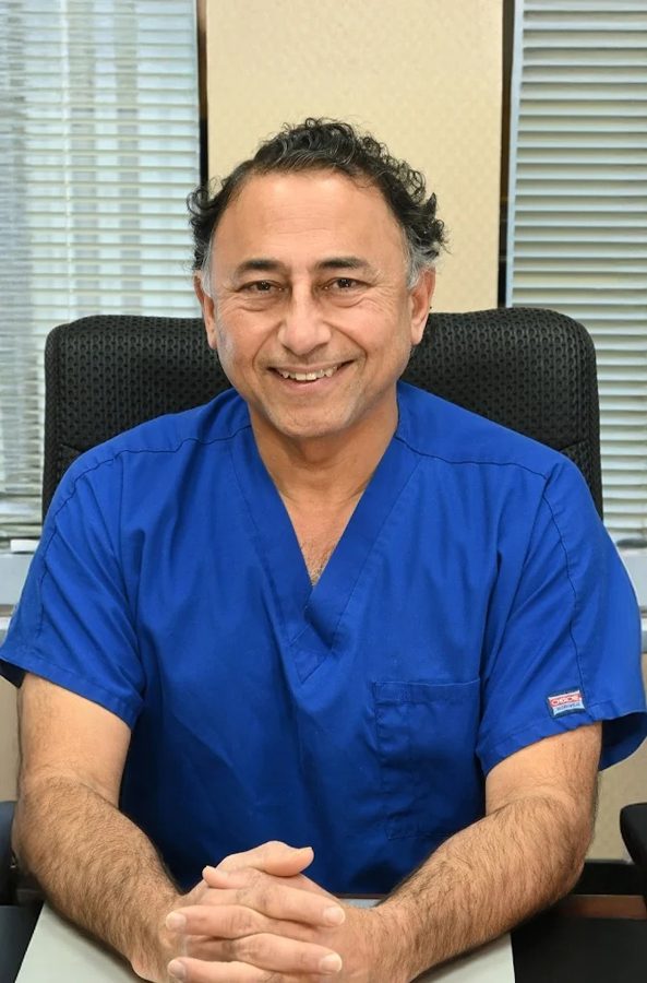 Dr. Rajesh Khanna in blue scrubs smiling at his desk at Khanna Vision Institute