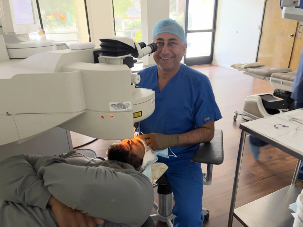 Dr. Rajesh Khanna in blue scrubs and a surgical cap smiling beside a laser system while a patient lies under it in a Khanna Vision Institute procedure room