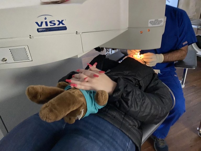 A patient holding a stuffed bear lies under a VISX excimer laser while a surgeon in blue scrubs works at Khanna Vision Institute